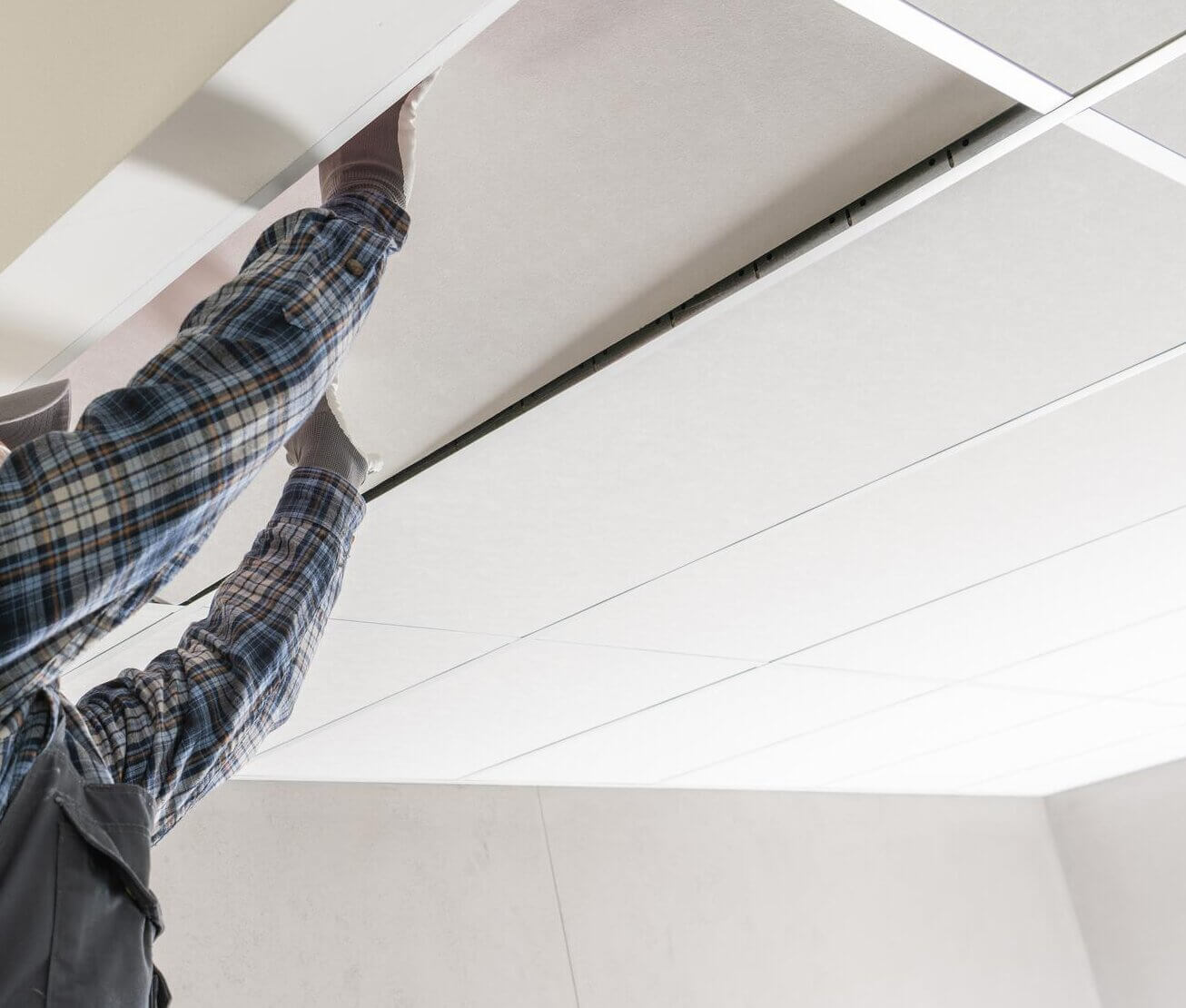 Mies asentaa valkoista alakattoa, jossa on metalliset tukirakenteet, käyttäen harmaita työkäsineitä ja siniruudullista paitaa. Huone on valoisa ja moderni.