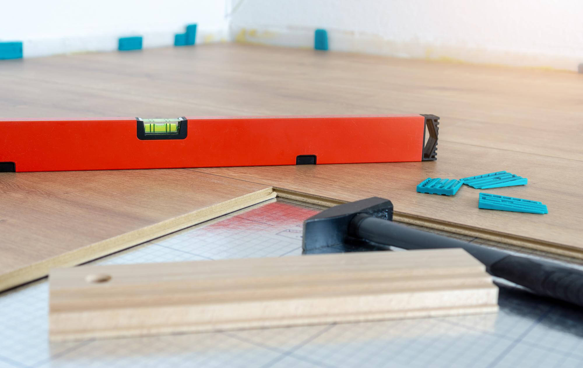 Lattiaremontti, jossa on vatupassi, vasara, lyöntipalikka ja sinisiä kiiloja laminaattilattialla, valoisassa huoneessa.