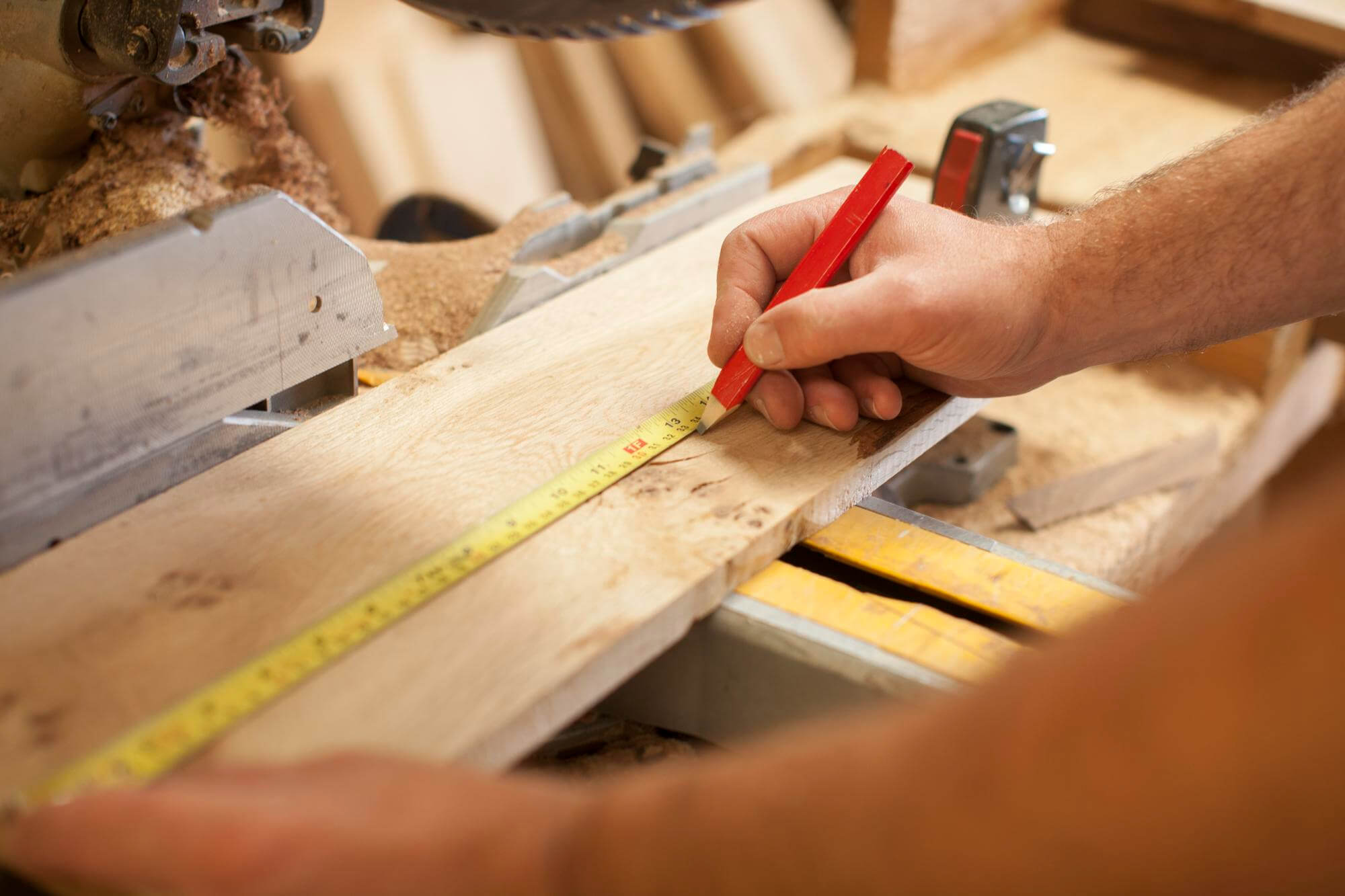Puuseppä mittaa puuta punaisella lyijykynällä ja mittanauhalla työpöydällä, jossa on puunpurua ja työkaluja. Kirkas valaistus.